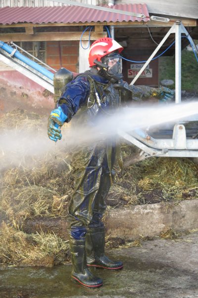 Einsatzkraft in Schutzanzug und Atemschutzmaske wird bei Tierrettung auf einem Bauernhof mit Hochdruckwasser dekontaminiert.